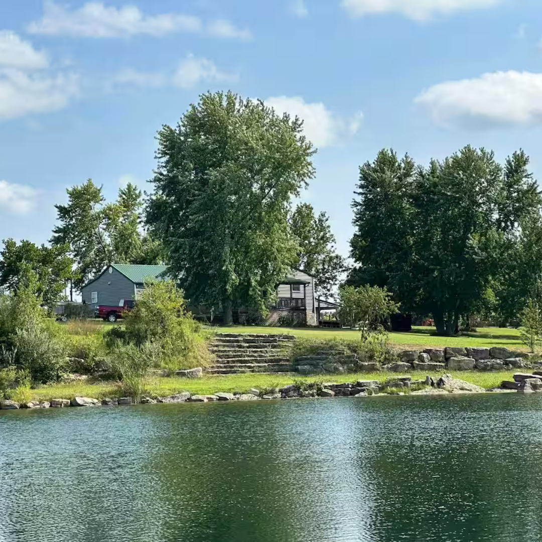 Lakeside view of Echevarria Hacienda, showcasing a cozy cabin surrounded by lush trees and stone steps leading to the water.